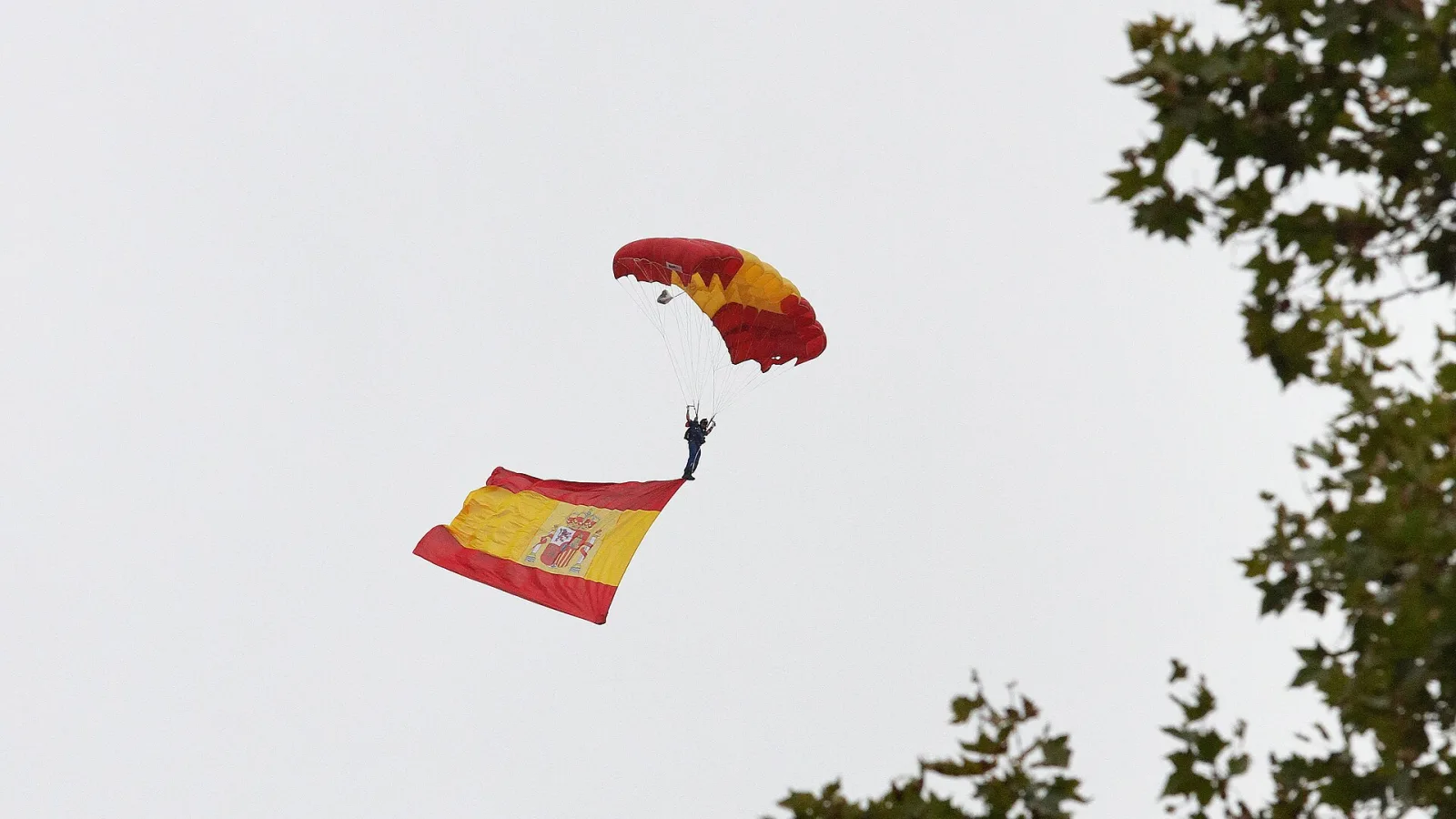 Paracaidista con un paracaídas rojo y amarillo que lleva colgada una gran bandera de España, recortado sobre un cielo blanco.