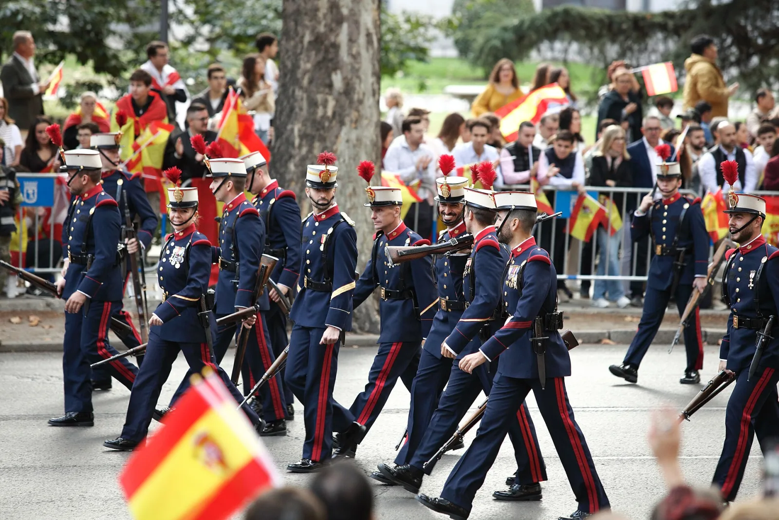 Militares con uniforme azul de gala y casco desfilan por una calle de Madrid; en primer plano, una bandera de España y público detrás de las vallas.