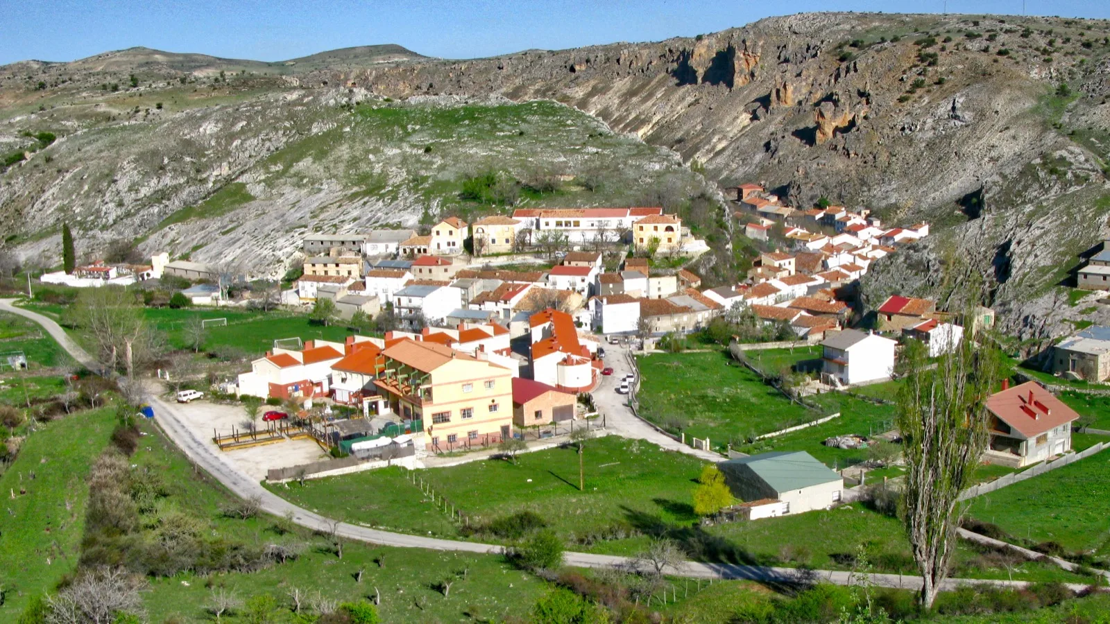 Vista desde lo alto de Pontones, en Santiago-Pontones: casas blancas de tejados rojos al pie de laderas de roca caliza, entre prados verdes y una carretera que rodea el pueblo.