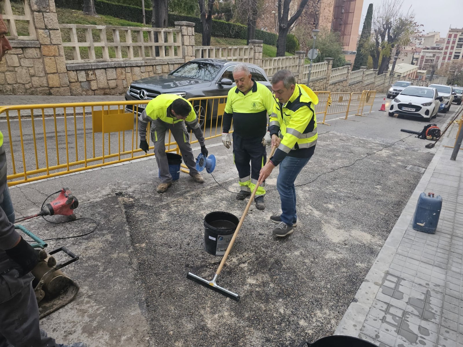 Tres operarios con chalecos reflectantes extienden material asfáltico con un rastrillo junto a vallas amarillas en una calle.