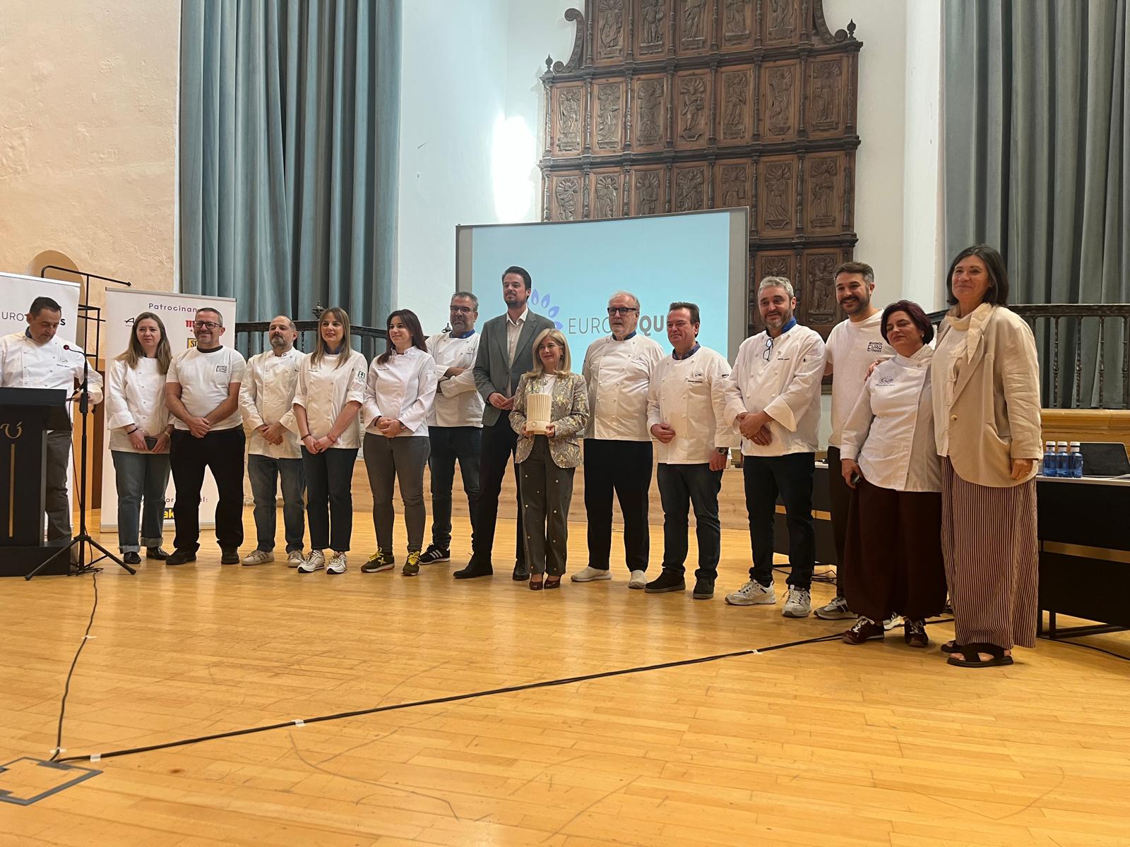 Un grupo de personas, muchas con chaquetilla blanca de cocinero, posa en fila sobre un escenario de madera ante una pantalla y un retablo tallado.