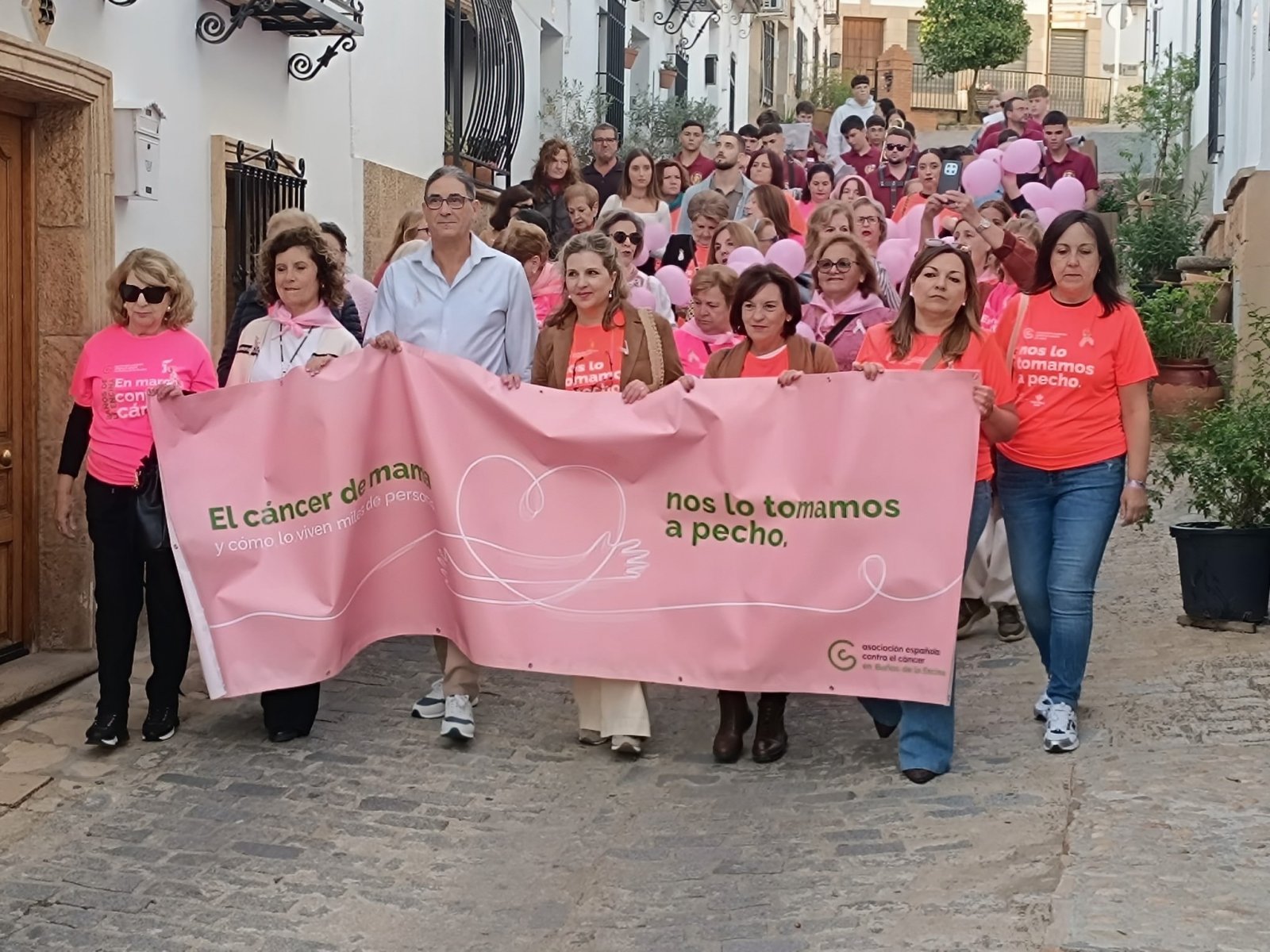 Una multitud con camisetas rosas y globos avanza por una calle empedrada portando una pancarta rosa contra el cáncer de mama.