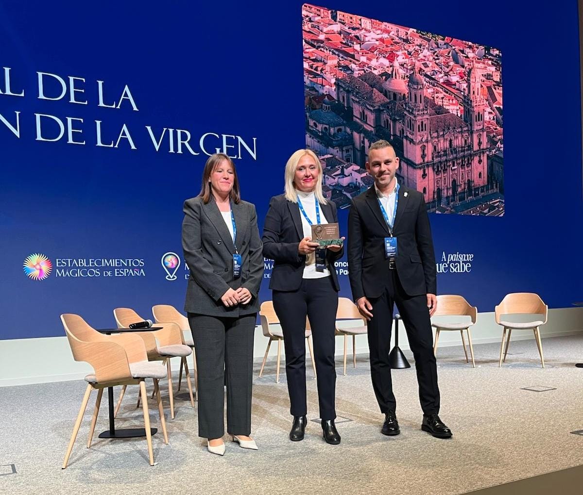 Tres personas trajeadas posan en un escenario ante una pantalla azul con un panel de una ciudad y sillas vacías.