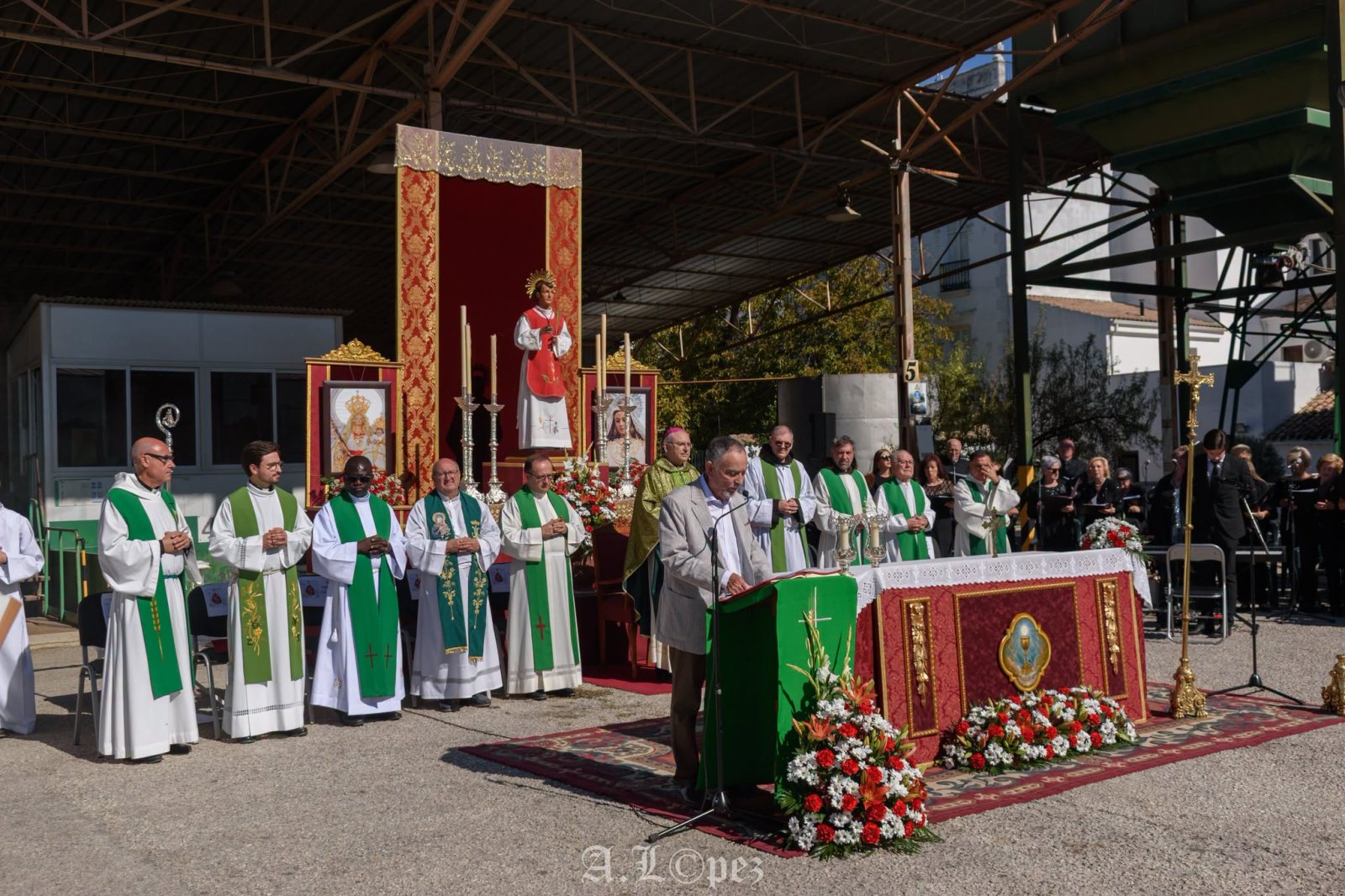 Un hombre con traje habla en un atril ante varios sacerdotes con vestiduras verdes y un altar con imágenes religiosas bajo una nave industrial.