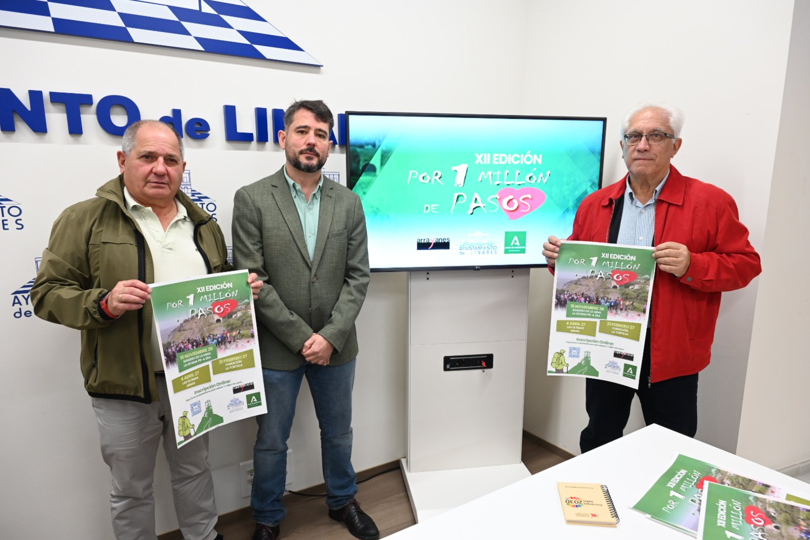 Tres hombres de pie sostienen carteles de una edición de senderismo ante una pantalla y un panel con letras azules.