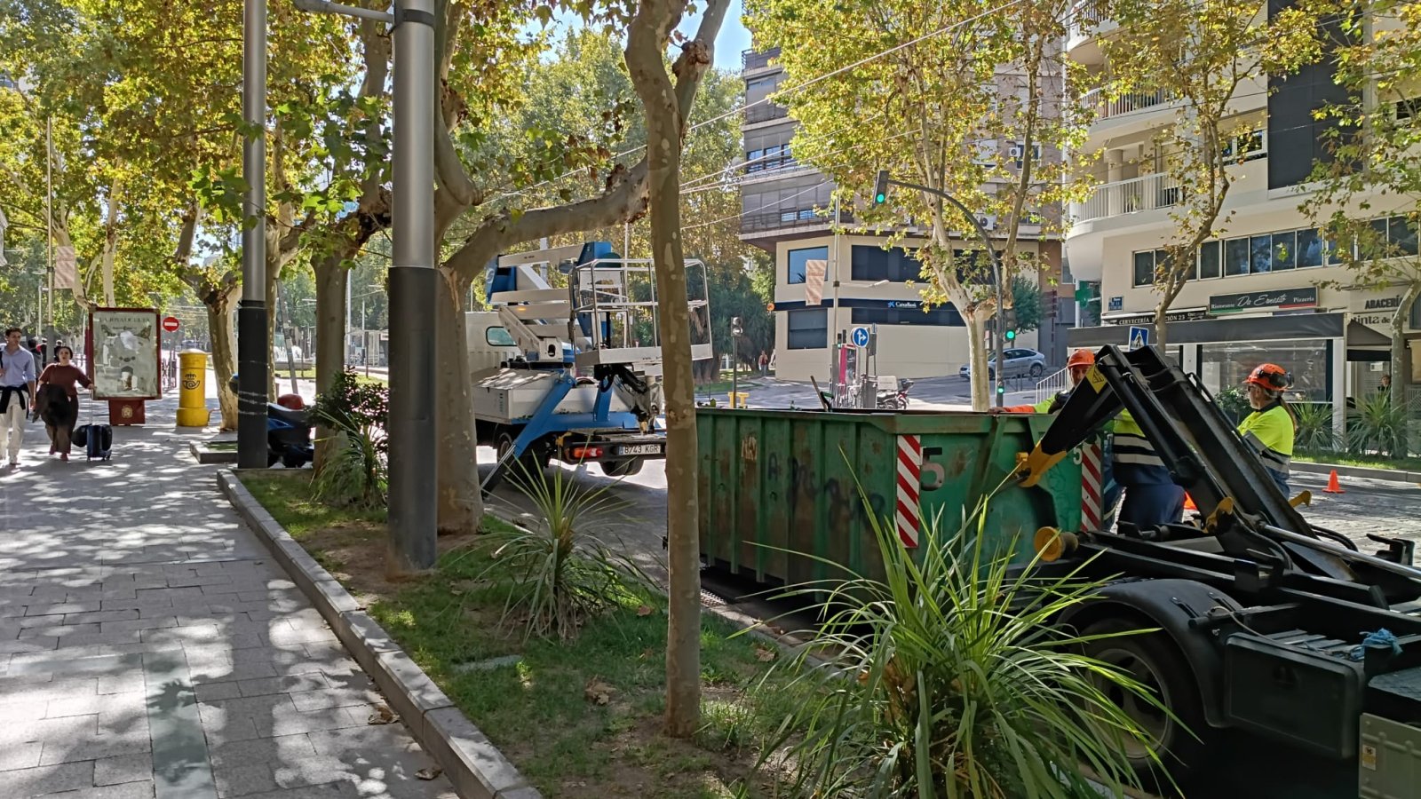 Un contenedor verde junto a un camión con plataforma elevadora y dos operarios con casco en una acera arbolada.
