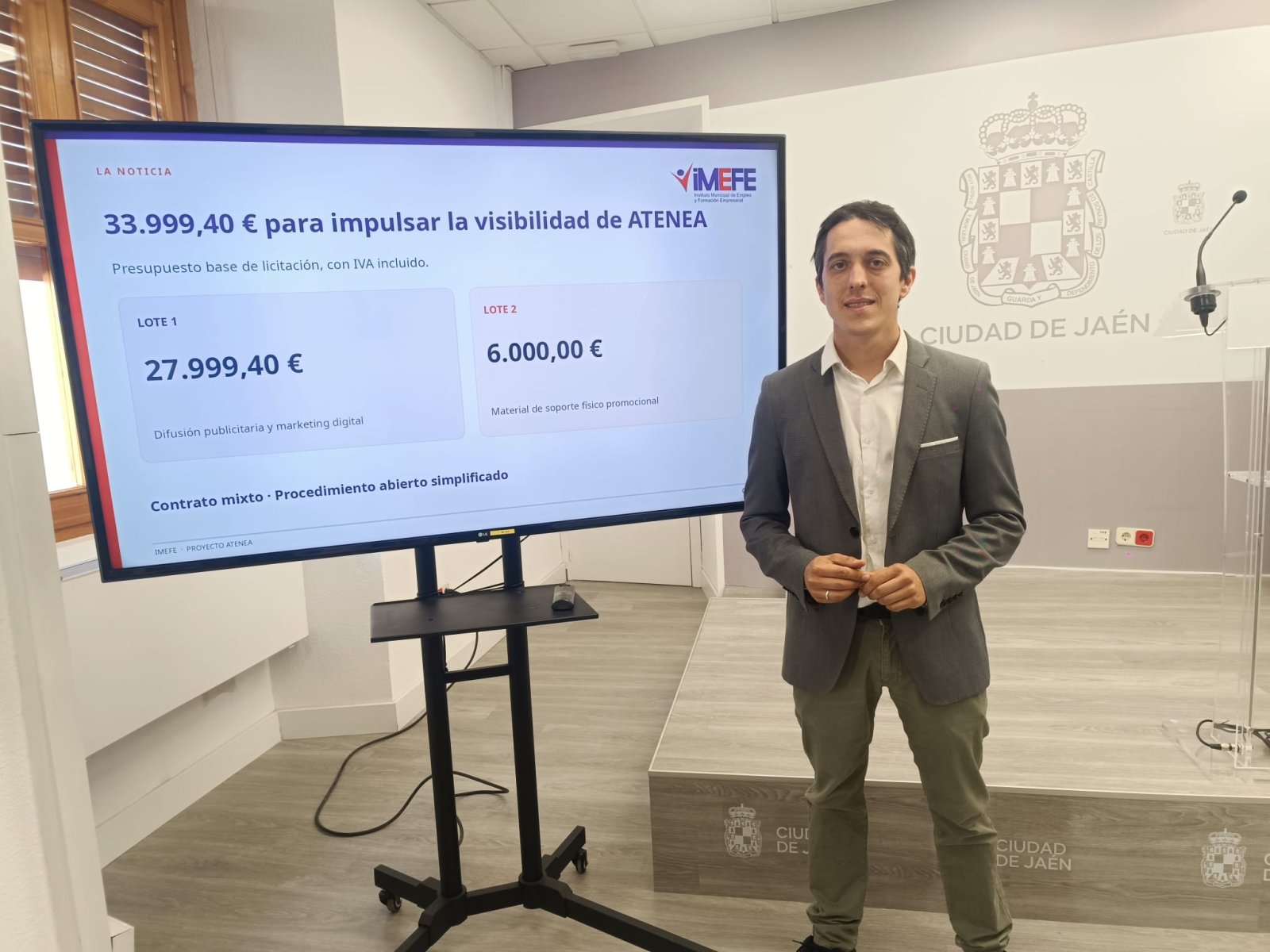 Un hombre trajeado posa junto a una pantalla grande con cifras de un presupuesto y el escudo de la ciudad de Jaén al fondo.