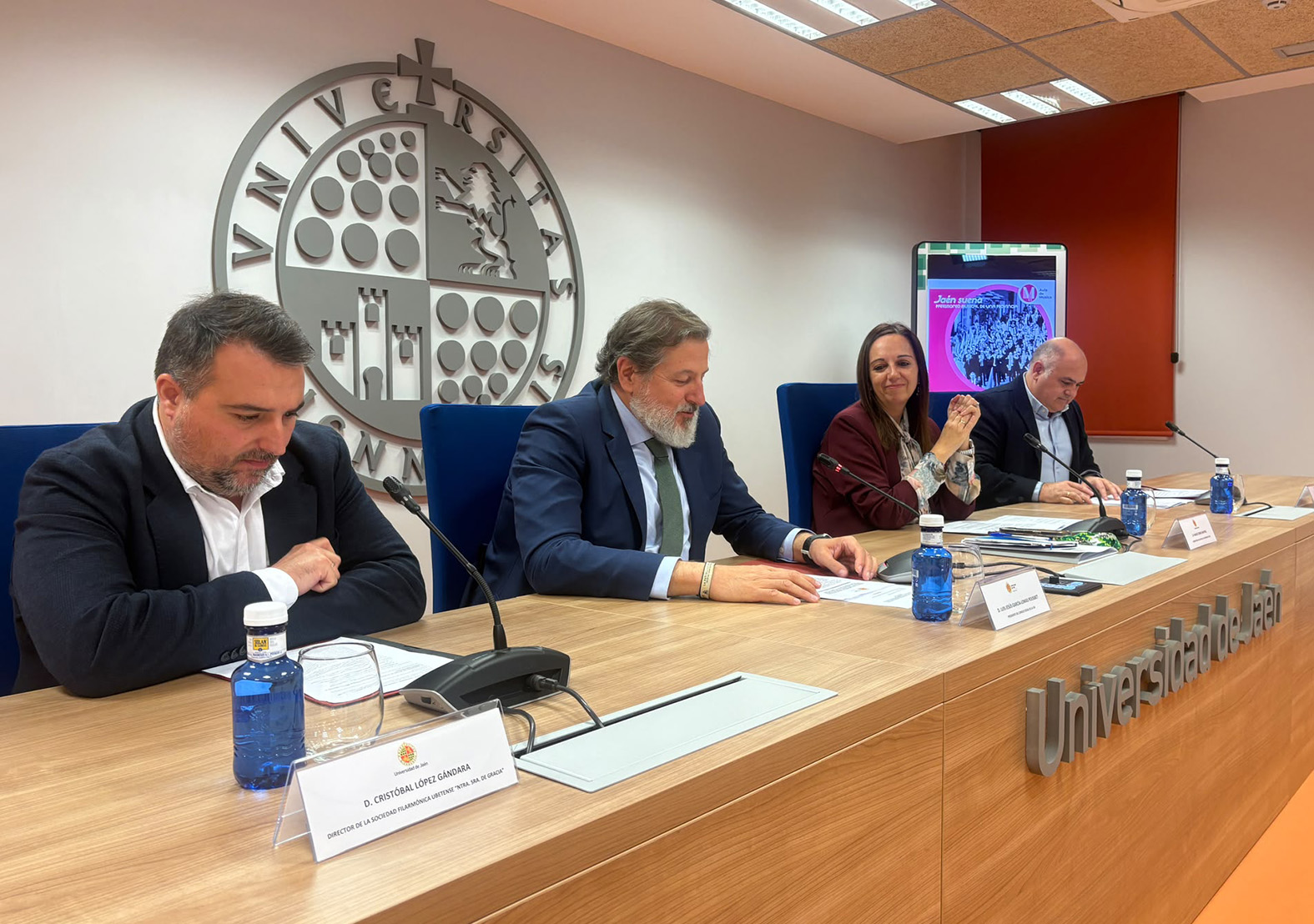 Cuatro personas con traje sentadas tras una mesa en una sala con el emblema circular de una universidad en la pared.