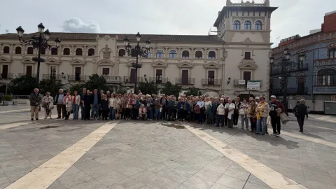 Un grupo numeroso de personas mayores posa en una plaza empedrada ante un edificio histórico con torre y farolas.