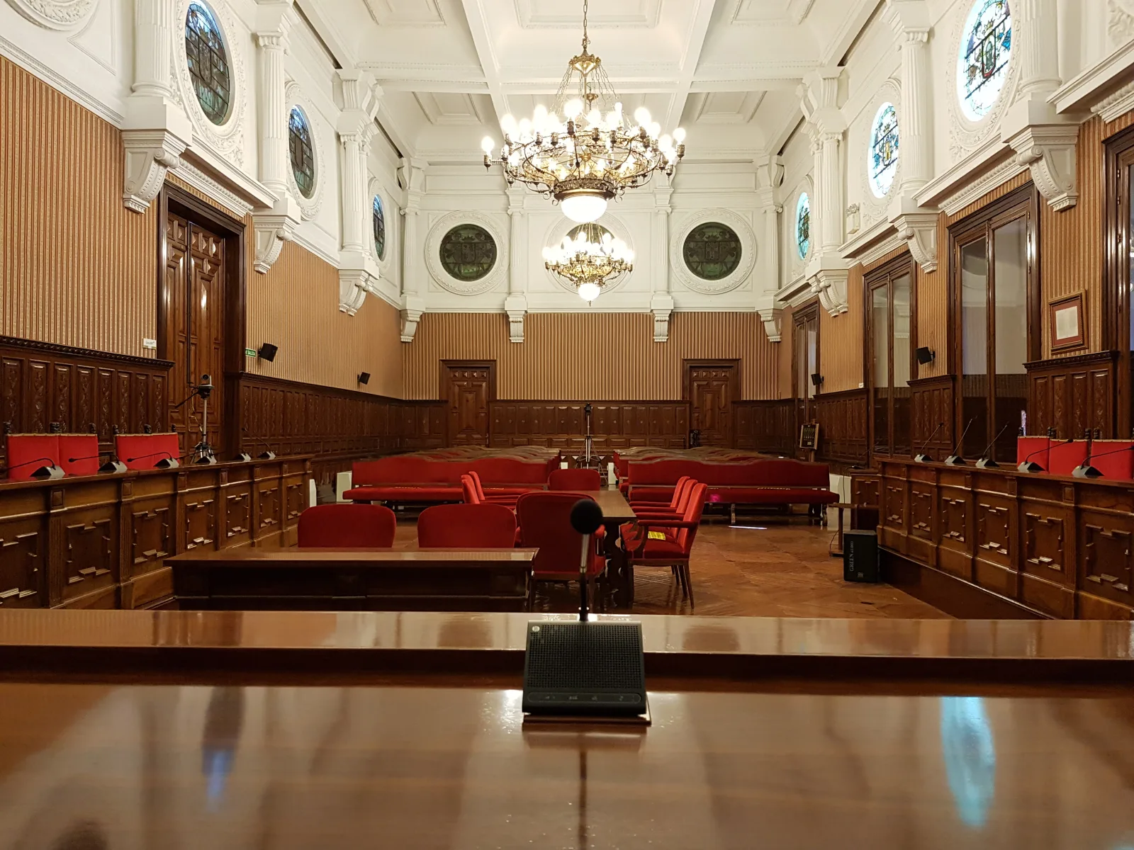 Sala de reuniones con paredes de madera, asientos rojos, micrófonos y lámparas de araña colgando del techo.