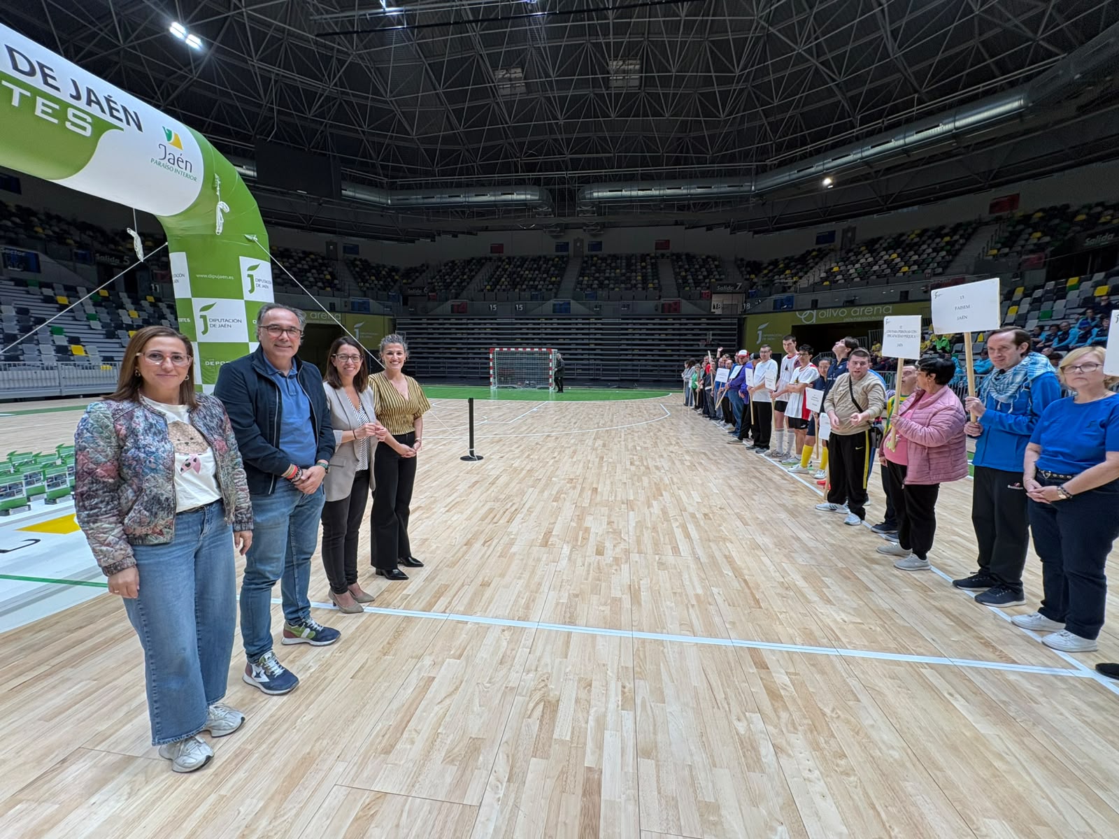 Cuatro personas posan en una cancha cubierta junto a un arco verde y una fila de participantes con carteles.