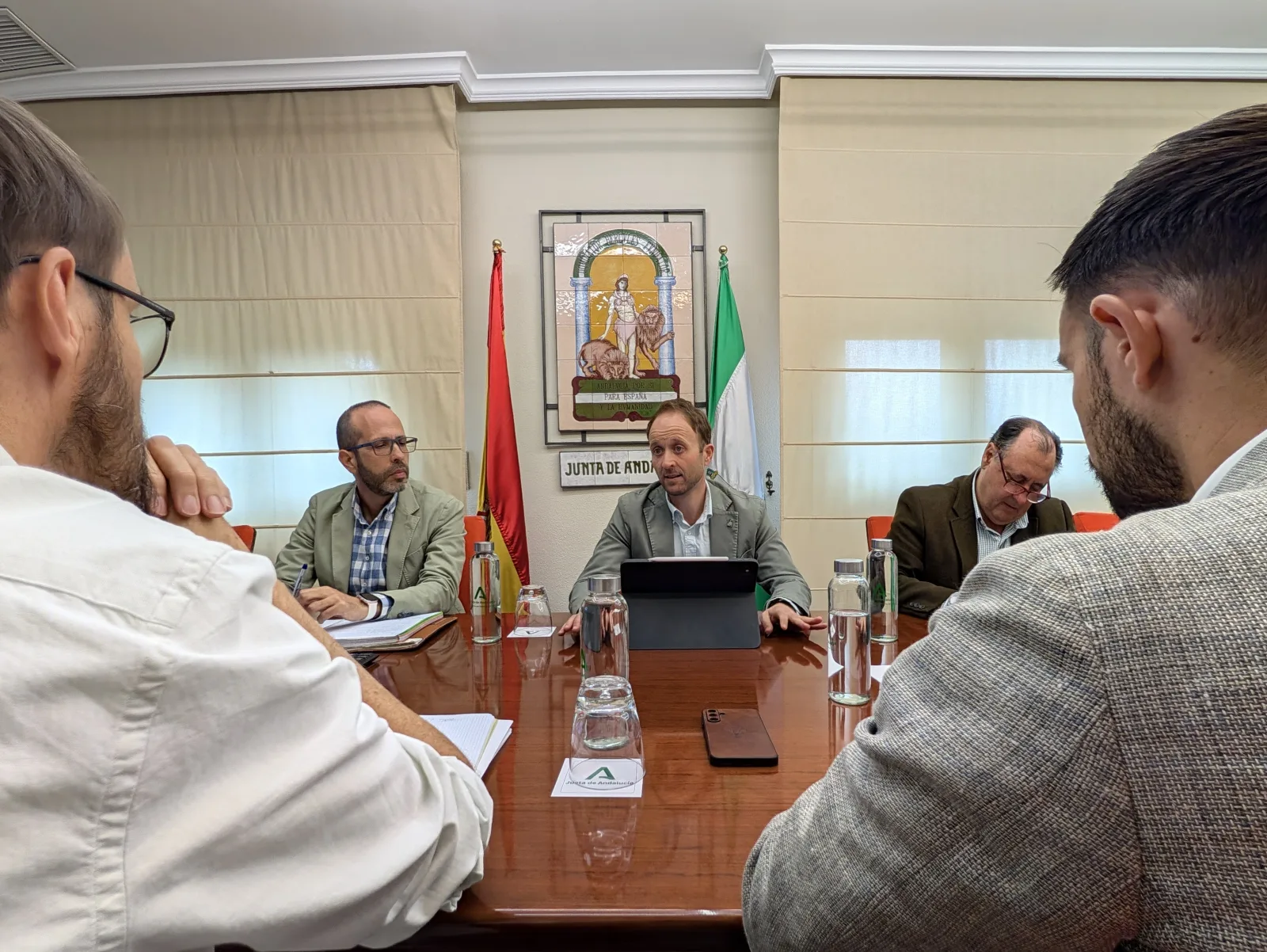 Cuatro hombres trajeados conversan alrededor de una mesa de reuniones con banderas de España y Andalucía al fondo.