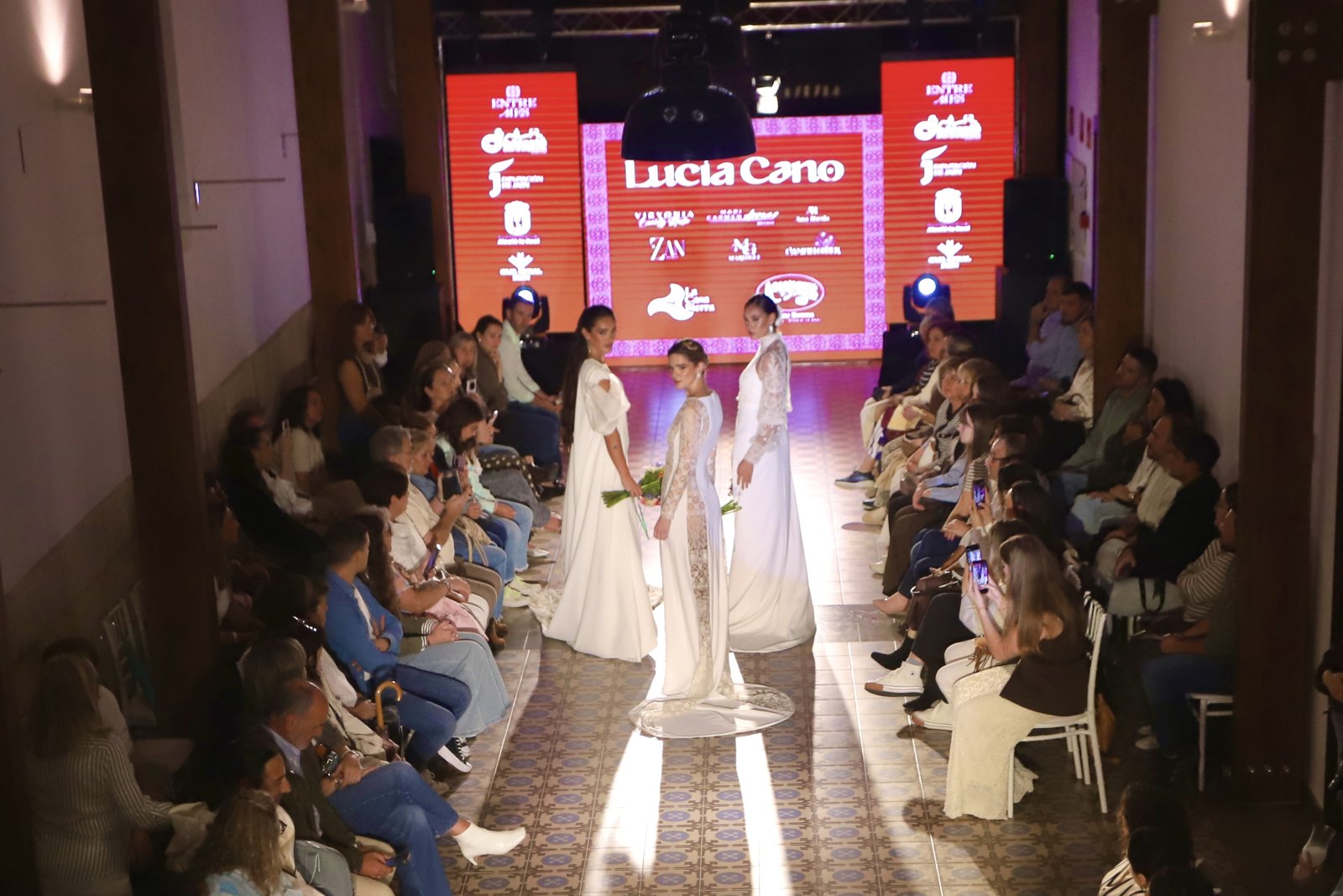 Tres modelos con vestidos blancos desfilan por un pasillo ante un público sentado a ambos lados y una pantalla roja al fondo.