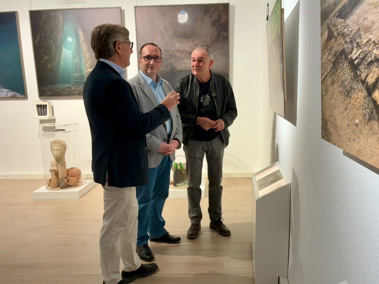 Tres hombres conversan de pie en una sala de museo con fotografías grandes en las paredes y una figura de piedra sobre una peana.