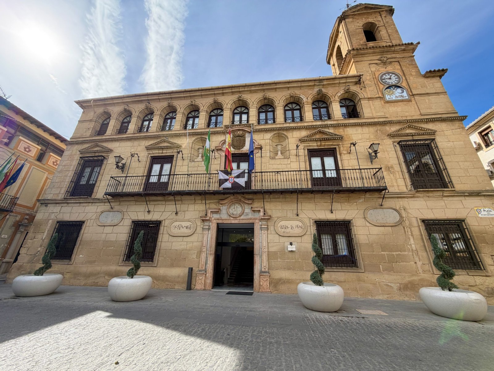 Edificio de piedra con balcón, banderas y torre con reloj, frente a una plaza con maceteros blancos.