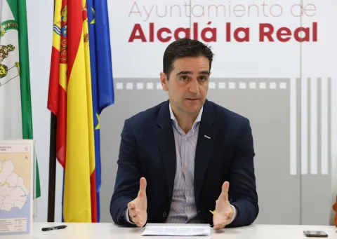 Un hombre trajeado gesticula sentado ante una mesa con papeles, banderas y el rótulo Ayuntamiento de Alcalá la Real.