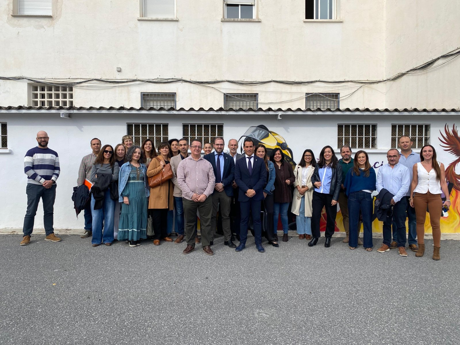 Un grupo de unas veinte personas posa de pie en una calle frente a la fachada blanca de un edificio con ventanas enrejadas y un mural.