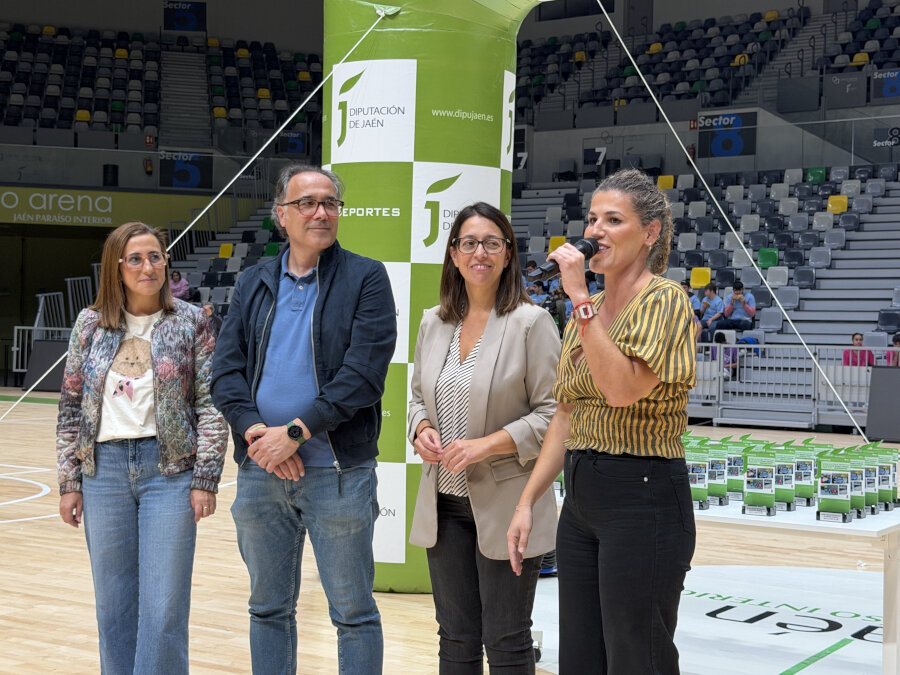 Cuatro personas de pie en una cancha cubierta, una con micrófono, junto a un hinchable verde y gradas con público.