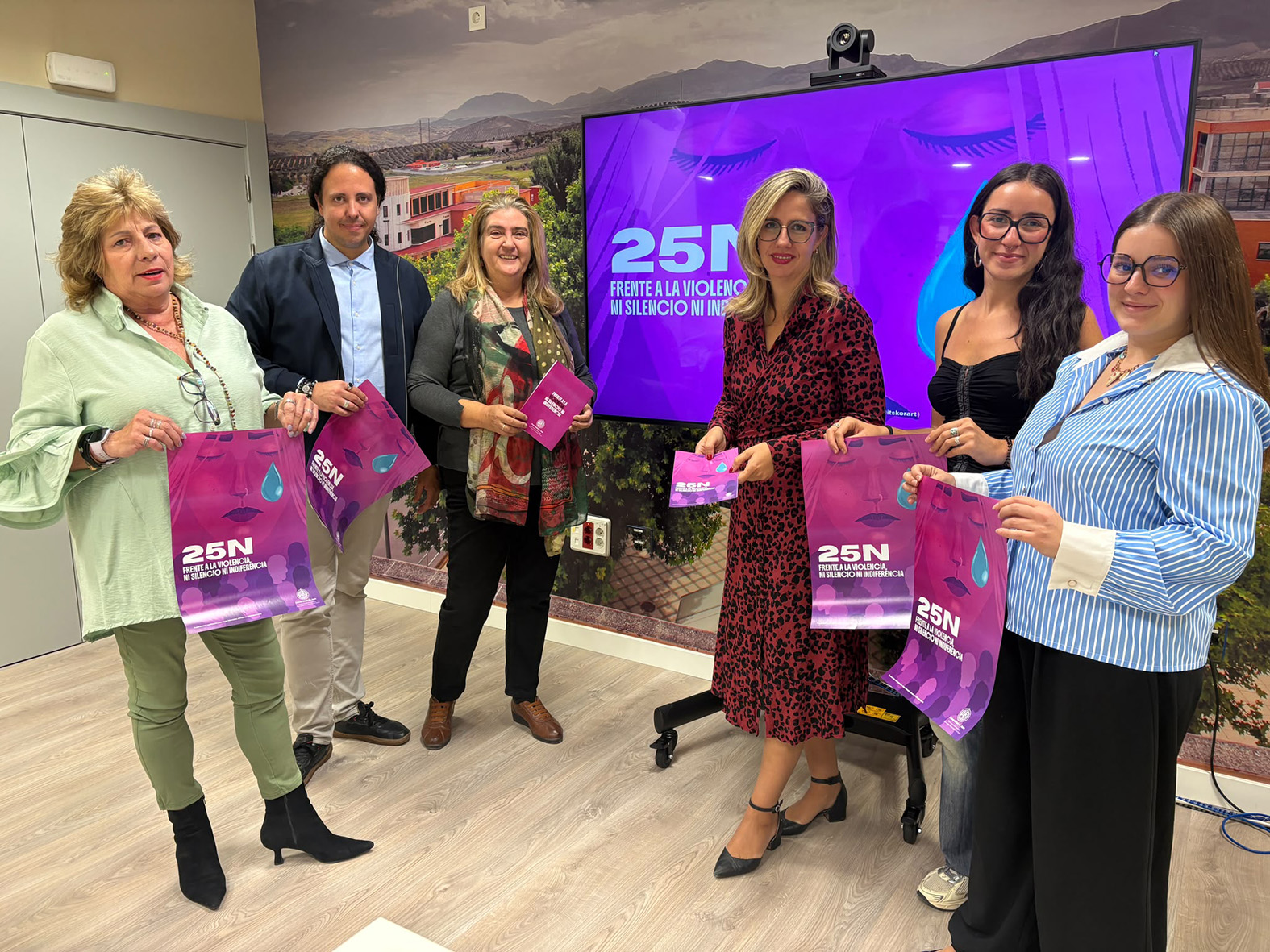 Seis personas de pie sostienen bolsas y folletos morados con el lema 25N ante una pantalla grande.