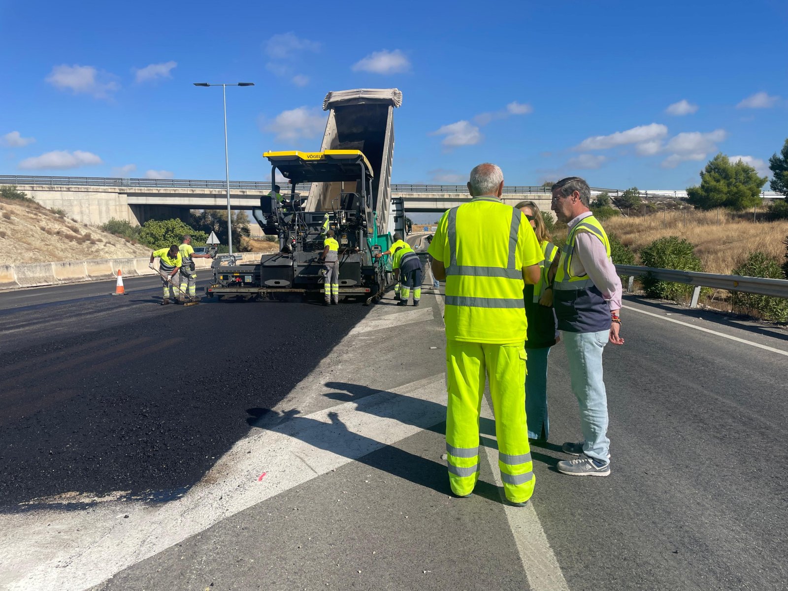 Operarios con chalecos reflectantes asfaltan una carretera junto a una extendedora, con un paso elevado al fondo.