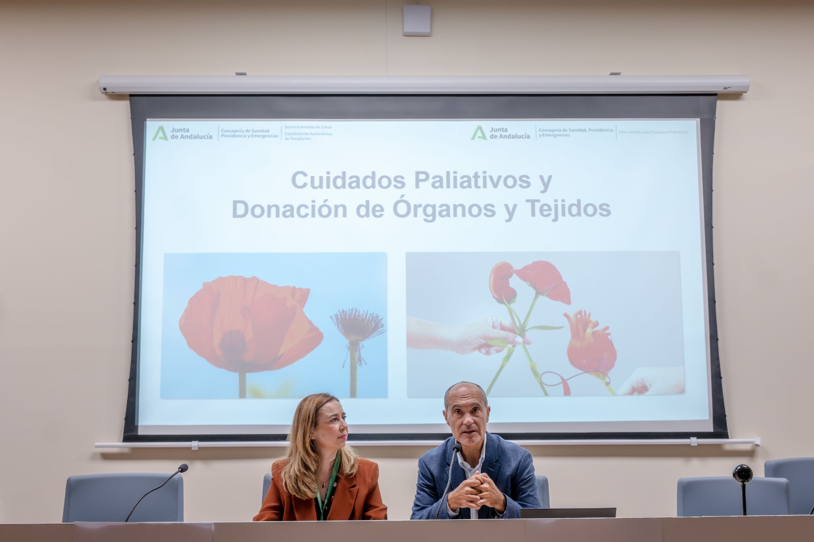 Un hombre y una mujer sentados tras una mesa ante una pantalla que proyecta una presentación sobre cuidados paliativos y donación.