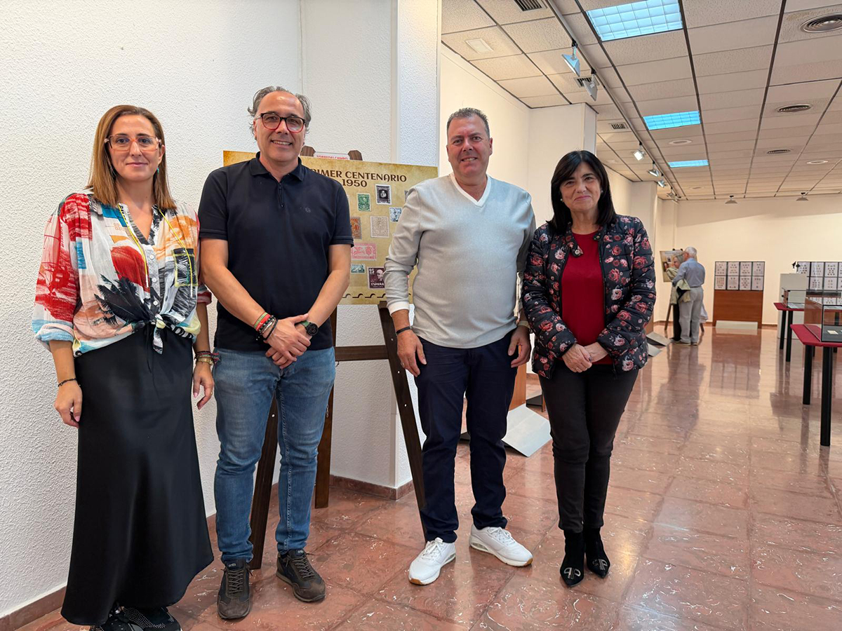 Cuatro personas de pie posan sonrientes junto a un panel con sellos en una sala de exposiciones con vitrinas al fondo.