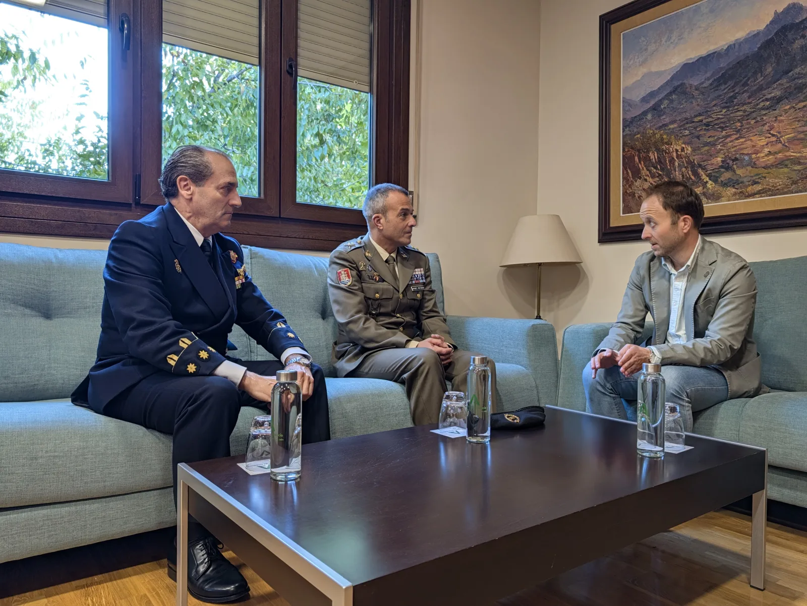 Tres hombres sentados en sofás de un despacho: dos con uniforme militar y uno de traje, con una mesa y botellas de agua delante.