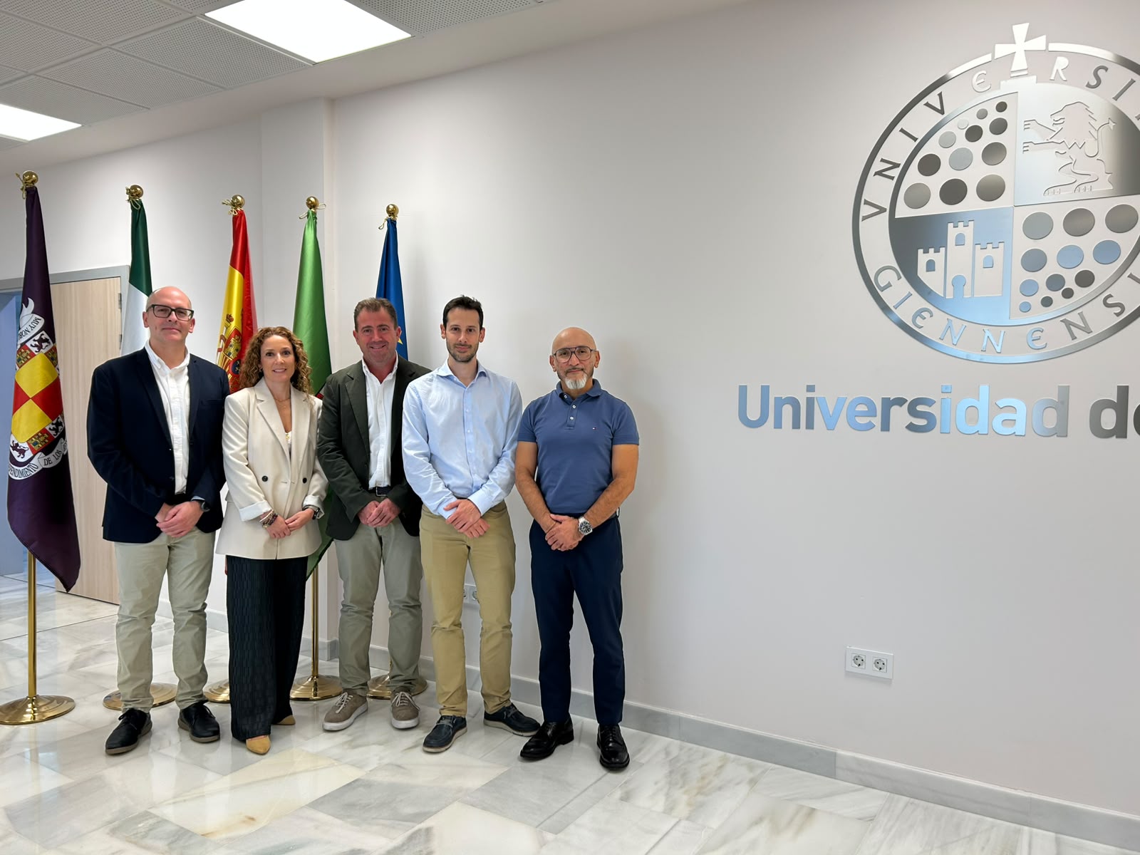 Cinco personas de pie posan junto a unas banderas ante un muro con el emblema y el rótulo de una universidad.