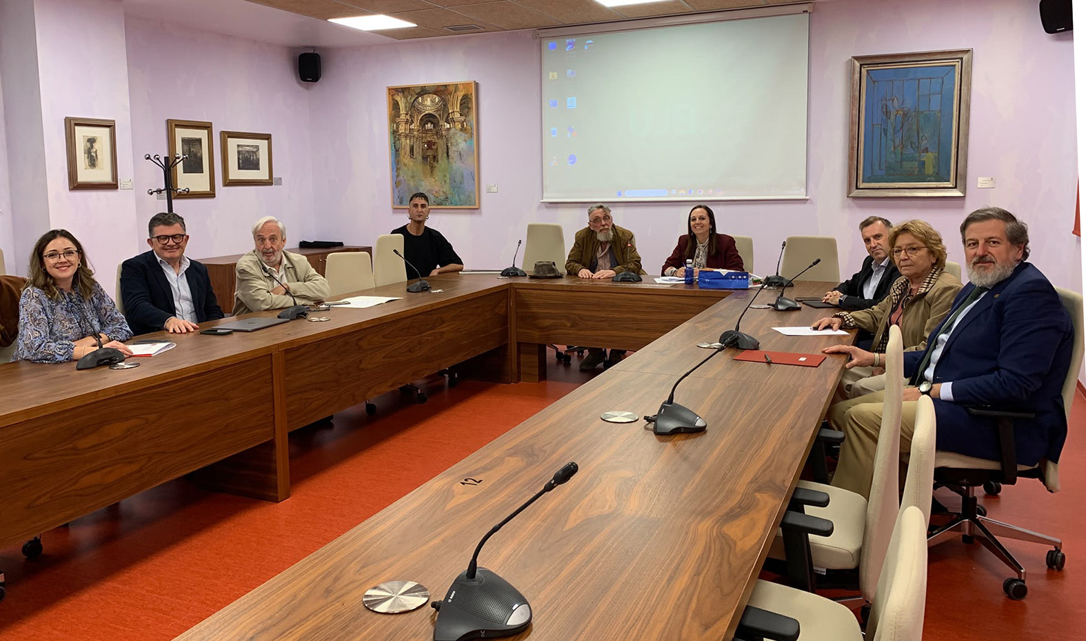 Ocho personas sentadas alrededor de una mesa de reuniones en forma de U, con micrófonos y una pantalla de proyección al fondo.