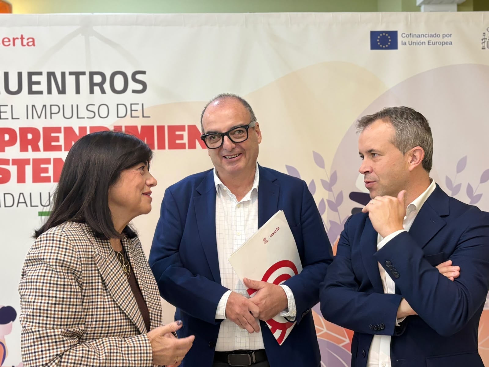 Tres personas conversando ante un cartel de un encuentro sobre emprendimiento, una de ellas con una carpeta.