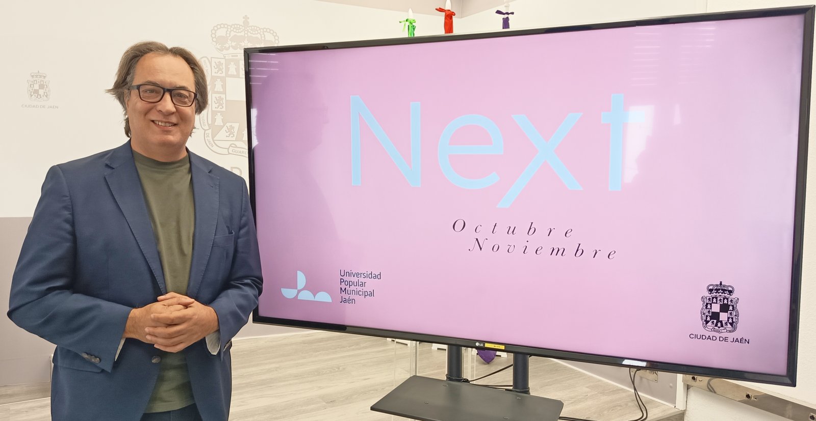 Un hombre con gafas y chaqueta azul posa junto a una pantalla grande que muestra el texto Next y Octubre Noviembre.