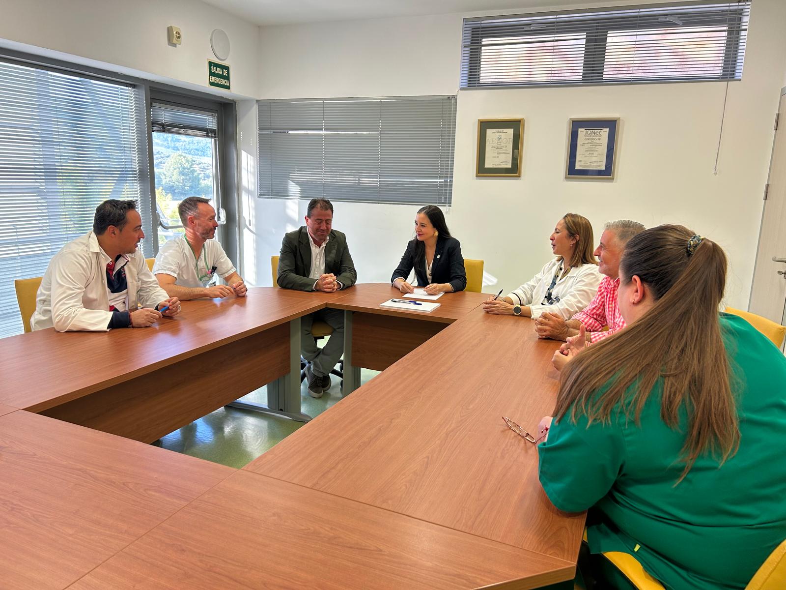 Siete personas sentadas alrededor de una mesa de reuniones en una sala con ventanas con persianas y un cartel de Salida de Emergencia.