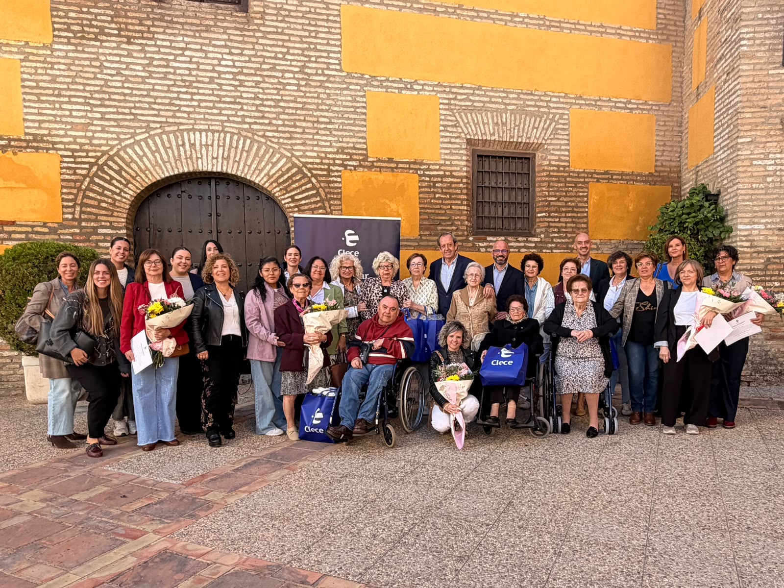 Un grupo numeroso de personas posa sonriente con ramos de flores y bolsas azules ante un edificio de ladrillo con arco y portón de madera.