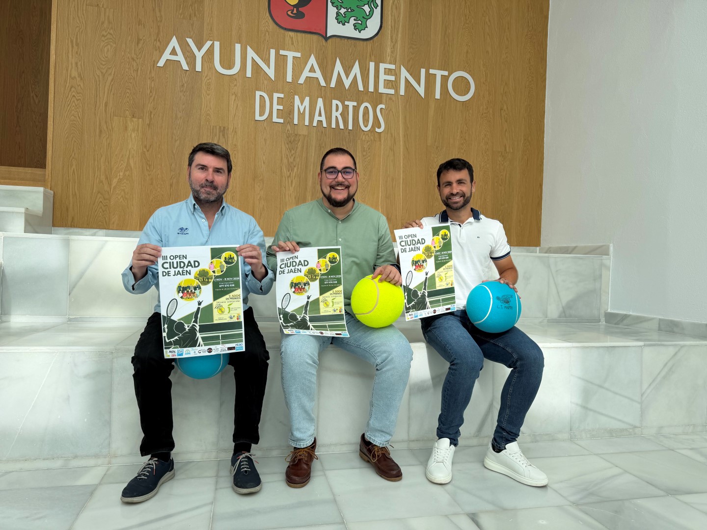 Tres hombres sentados en unos escalones sostienen carteles del III Open Ciudad de Jaén y pelotas de tenis.