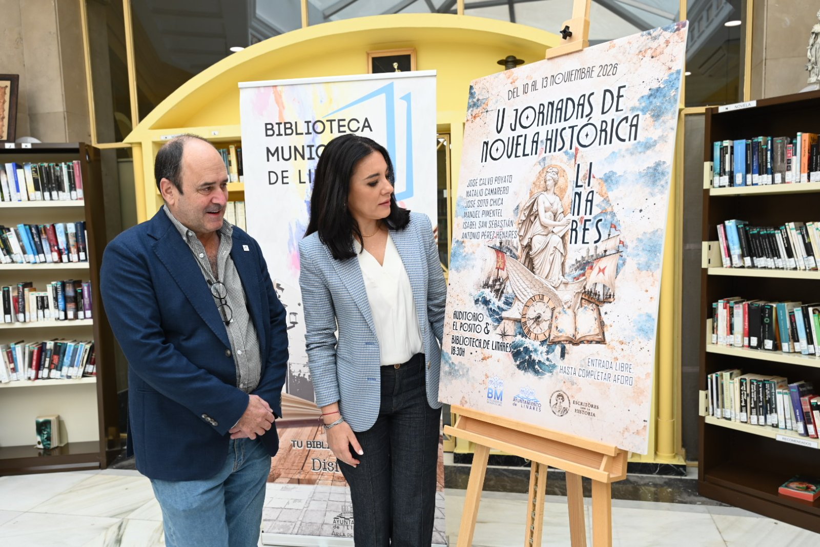 Un hombre y una mujer junto a un cartel de unas jornadas de novela histórica, entre estanterías con libros.