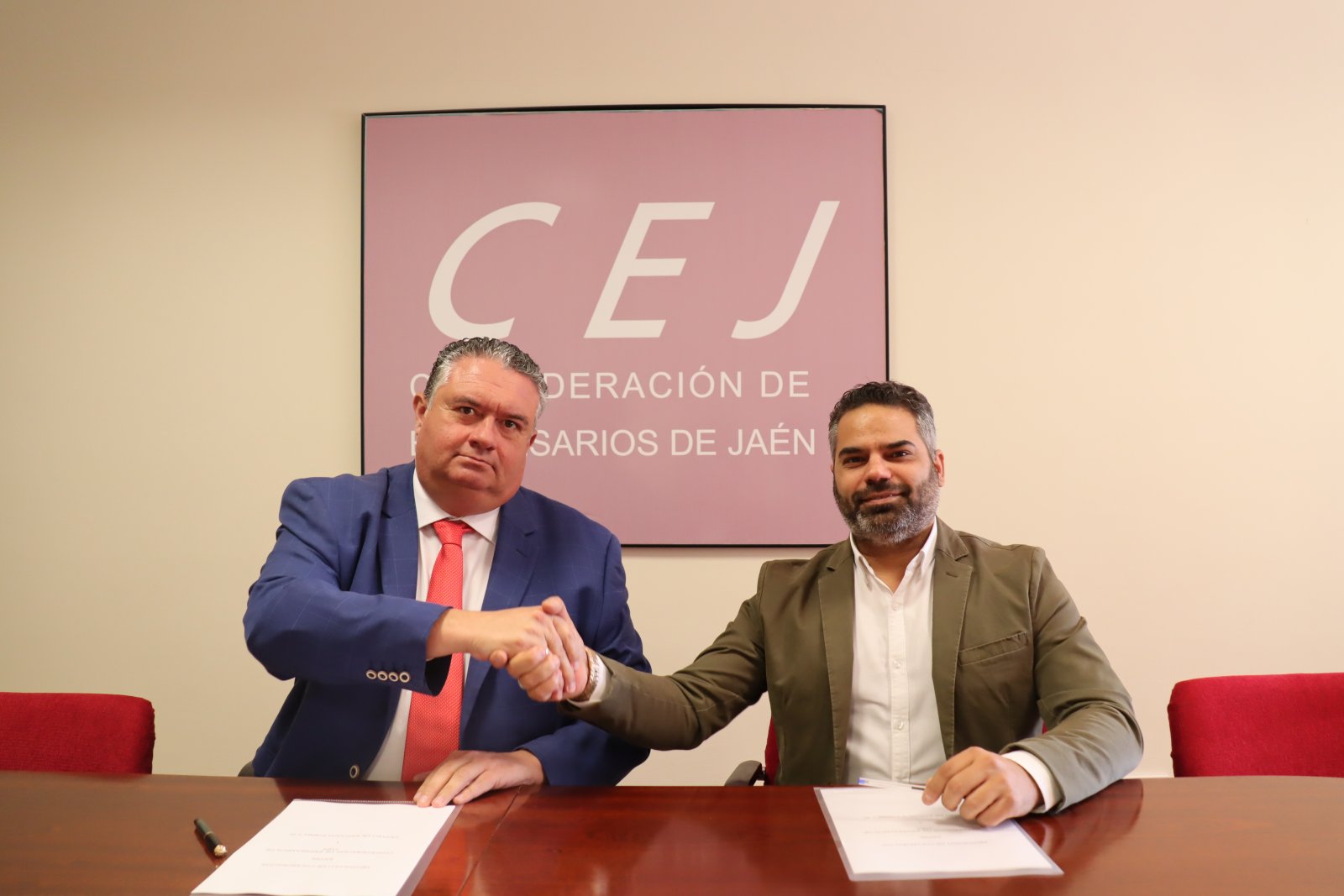 Dos hombres trajeados se dan la mano sentados ante una mesa con documentos, con un cartel de la CEJ al fondo.