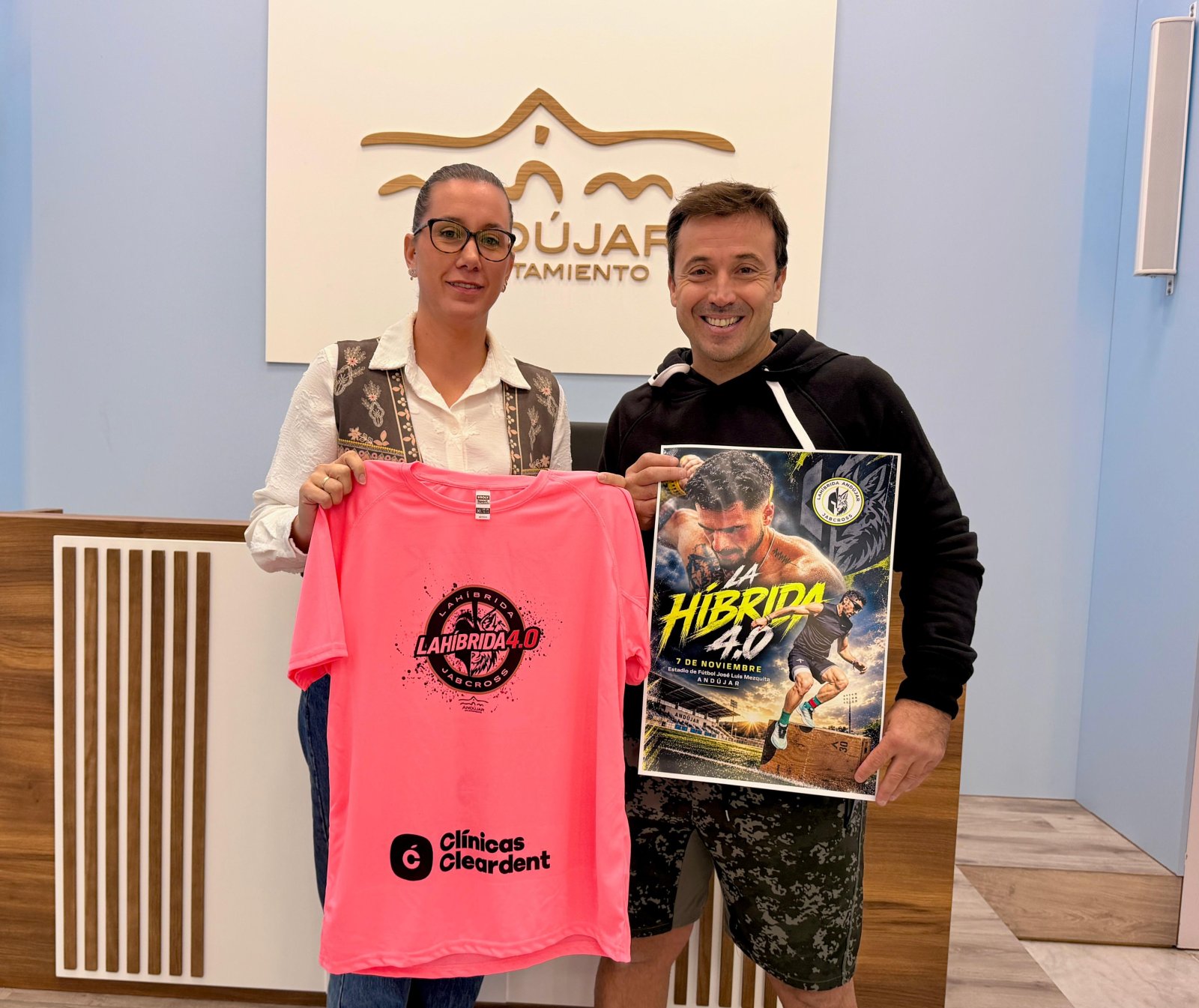 Un hombre y una mujer sostienen una camiseta rosa y un cartel de una carrera ante un panel con un logotipo municipal.