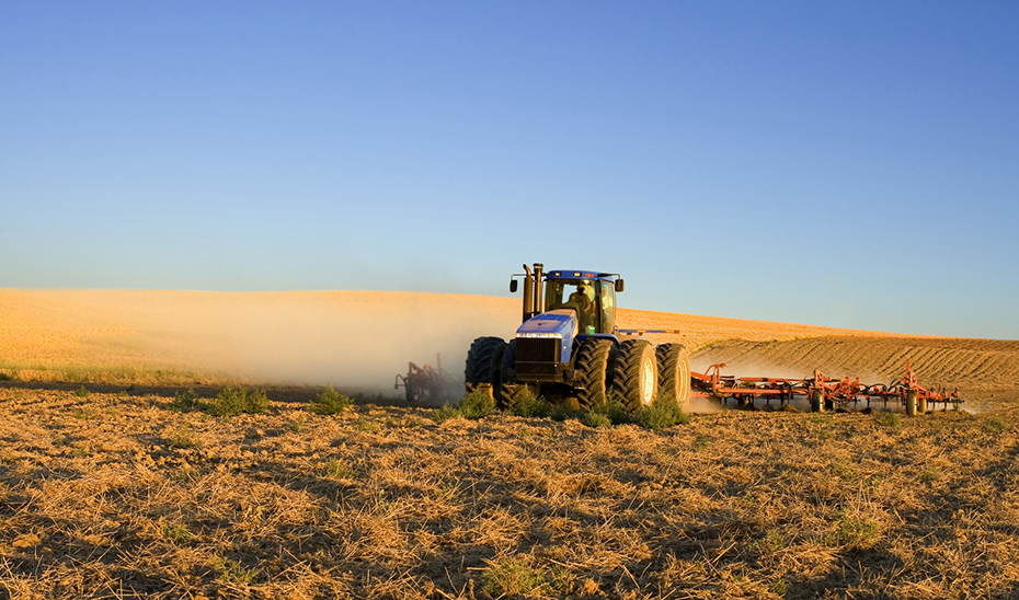 Un tractor azul con apero de labranza trabaja en un campo de rastrojos, levantando polvo bajo un cielo despejado.