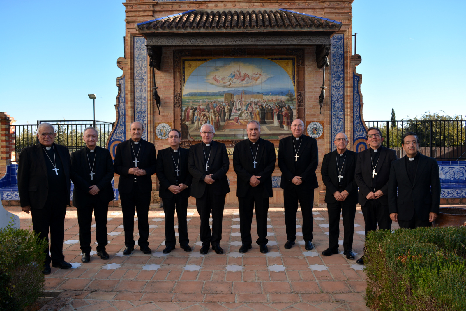 Diez hombres con traje negro y cruz pectoral posan en fila ante un retablo cerámico de azulejos con una escena religiosa.