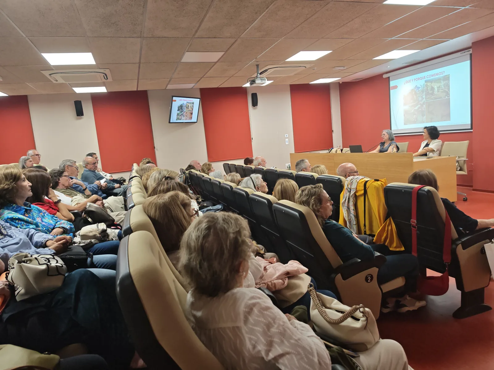 Público sentado en un aula escuchando a dos mujeres tras una mesa junto a una pantalla de proyección.