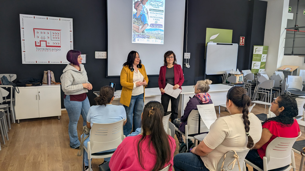 Tres mujeres de pie hablan ante varias personas sentadas en sillas, en un aula con proyector y carteles en la pared.