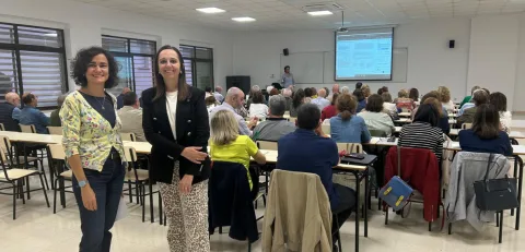 Dos mujeres de pie sonríen en primer plano ante un aula con alumnos sentados y una pantalla de proyección al fondo.