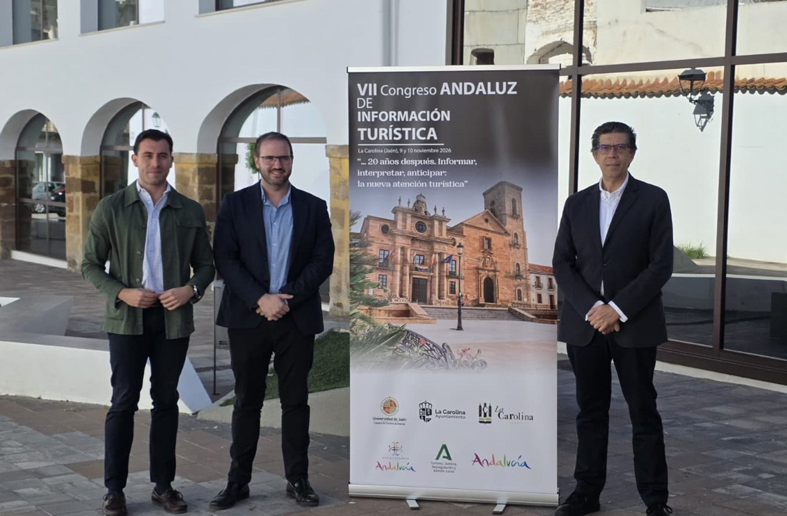 Tres hombres trajeados posan junto a un cartel vertical sobre un congreso andaluz de información turística ante un edificio con arcos.