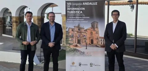 Tres hombres trajeados posan junto a un cartel vertical sobre un congreso de información turística ante un edificio con arcos.