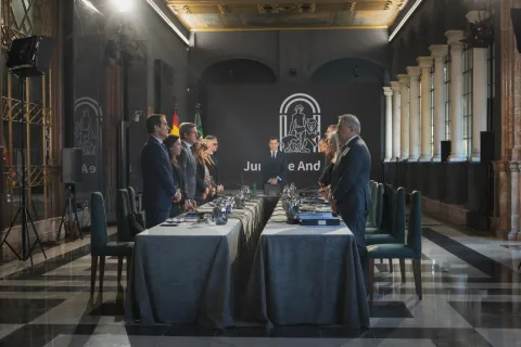 Varias personas trajeadas de pie junto a una mesa larga en una sala con columnas y un emblema al fondo.