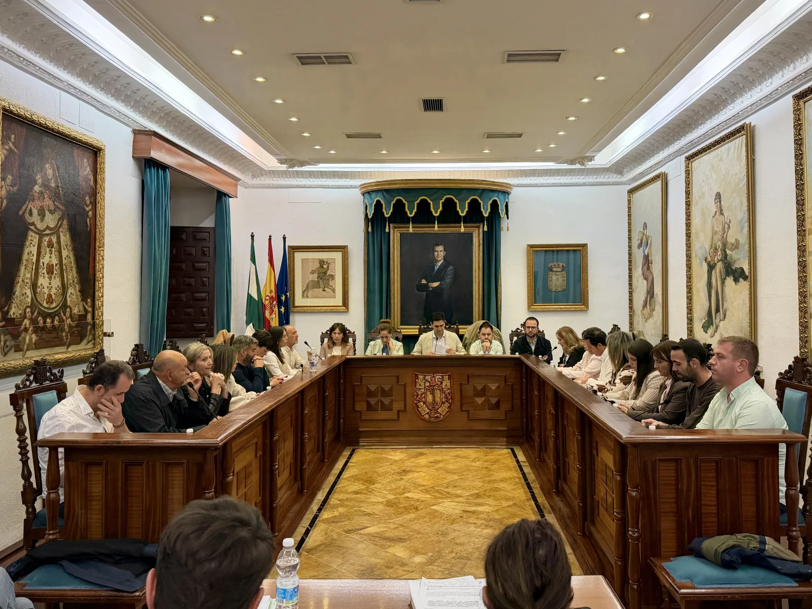 Varias personas sentadas alrededor de una mesa de madera en U en una sala con retratos, banderas y escudo.