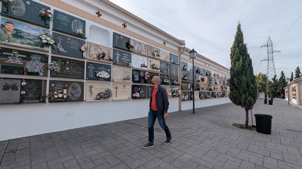 Un hombre camina junto a una larga pared blanca con nichos y placas florales de un cementerio.
